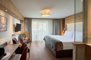 A modern hotel room with a bed, a desk with a chair, a television, and a large window with curtains. - DOUBLE ROOM BERGKRISTALL - 5* Wellness & Sporthotel Jagdhof (Röhrnbach)