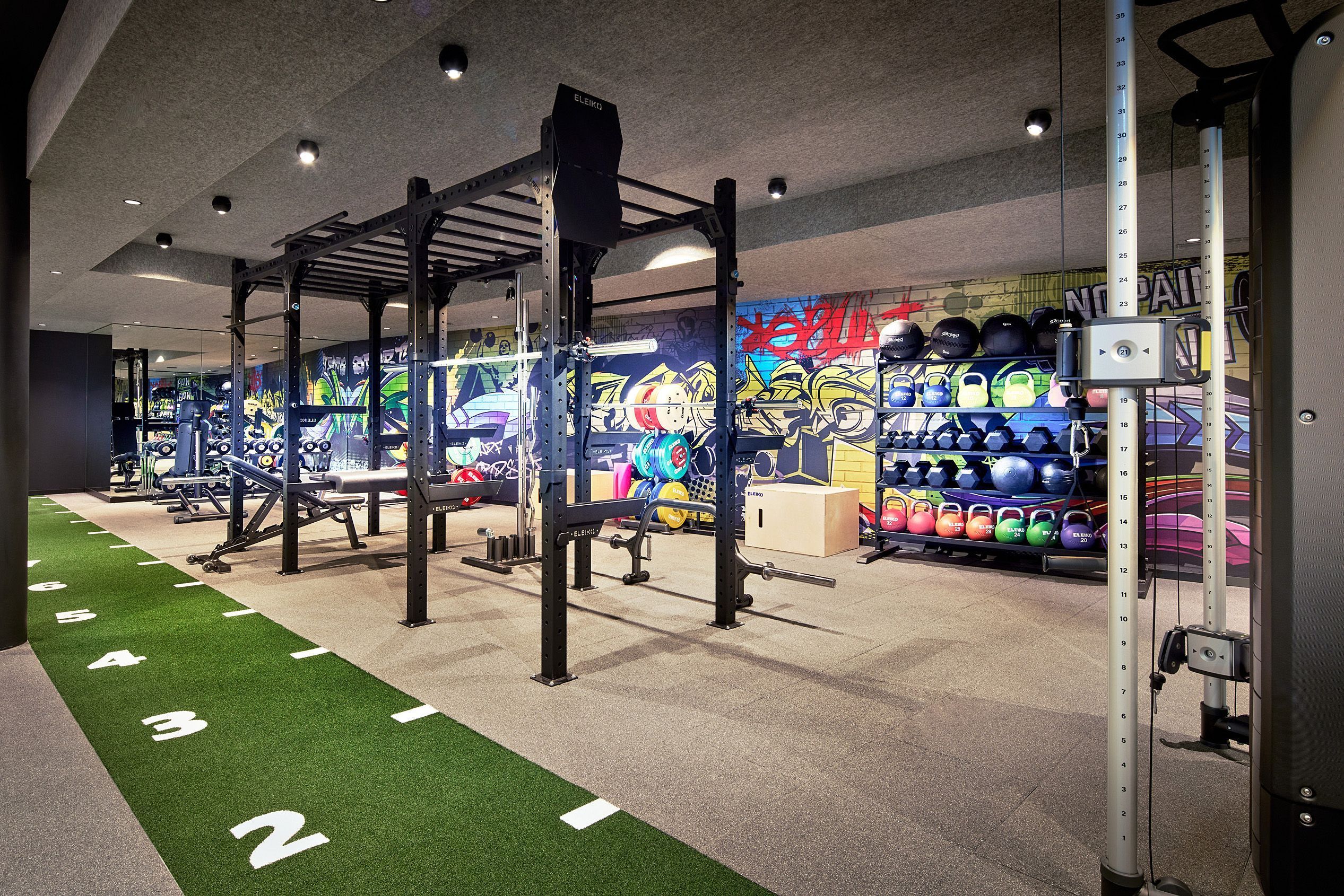Et fitnesscenter med vægttræningsudstyr, en grøn løbedekke og en væg med farverige graffiti.