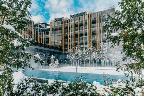 A modern building with a glass facade, surrounded by snow-covered trees and a pool.