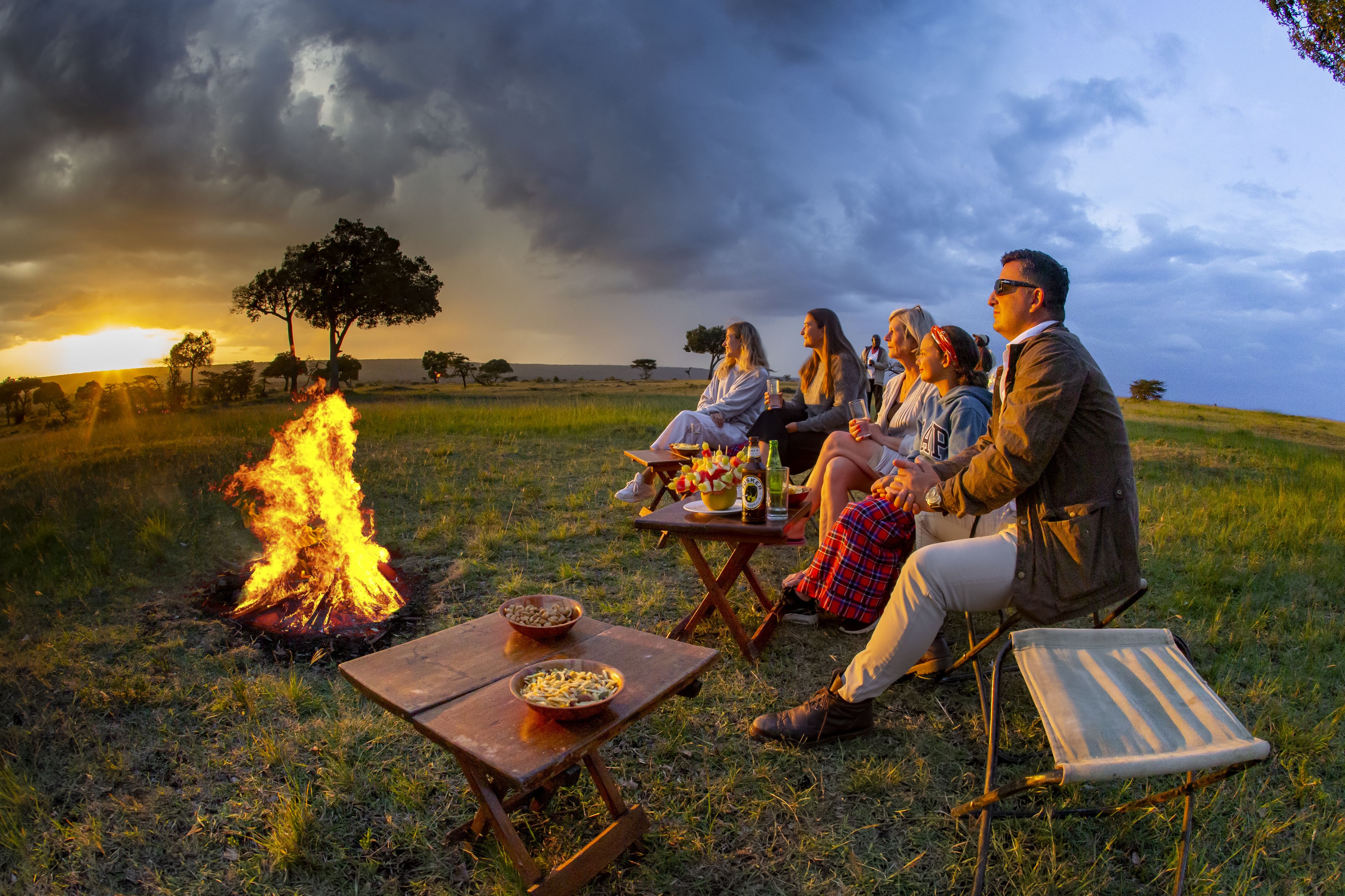 A group of people sitting around a campfire in an open field with a sunset in the background.