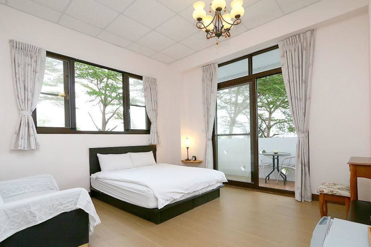 Un dormitorio con cama, escritorio y silla. Hay ventanas con cortinas, un candelabro y vistas a árboles desde el exterior.