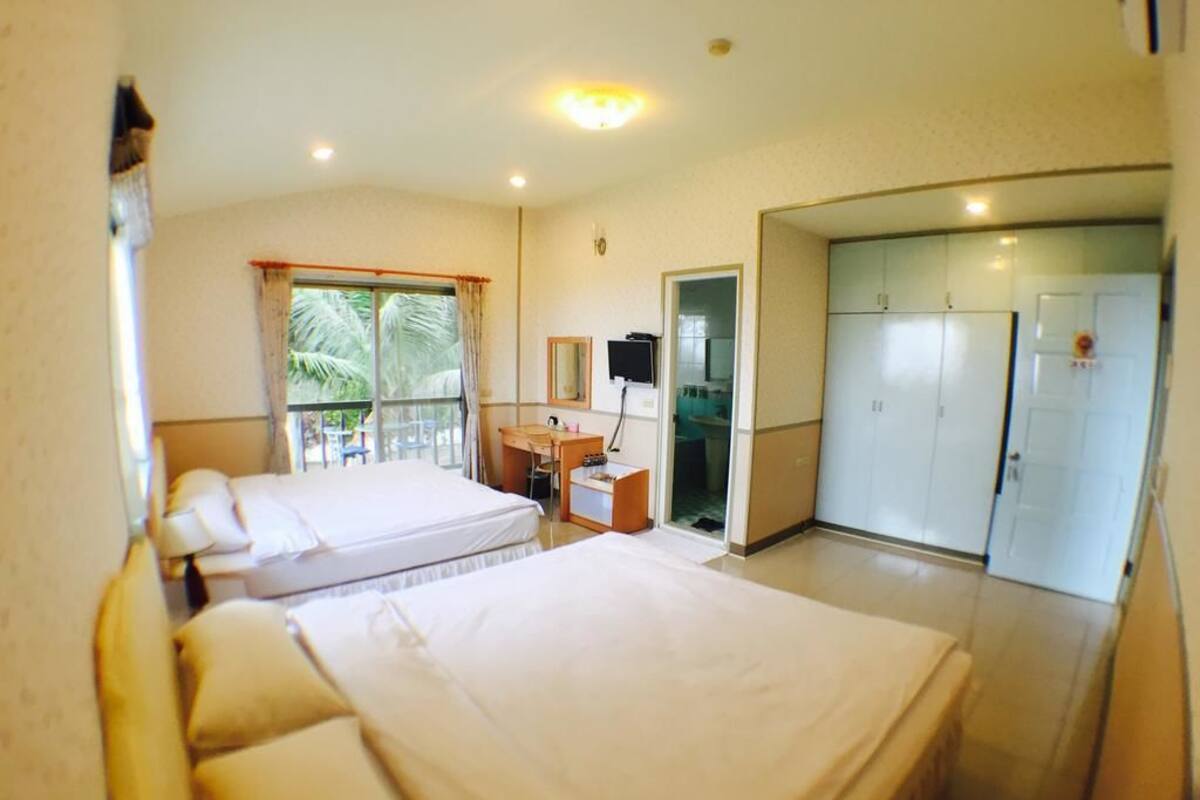 Habitación de hotel con dos camas, un escritorio, un armario y un balcón con vista a palmeras.