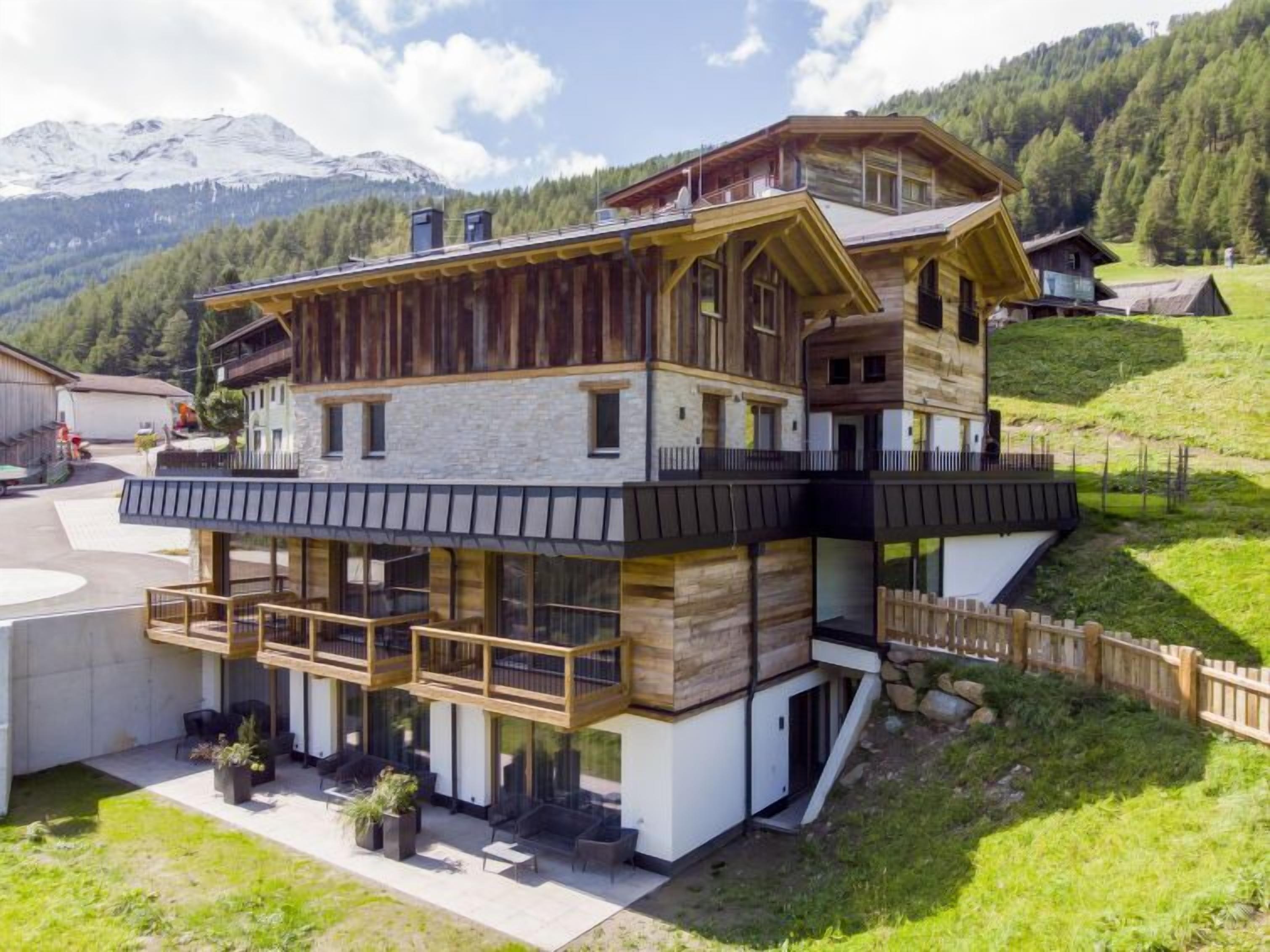 A multi-story wooden chalet with balconies, situated on a hillside with a mountain backdrop.