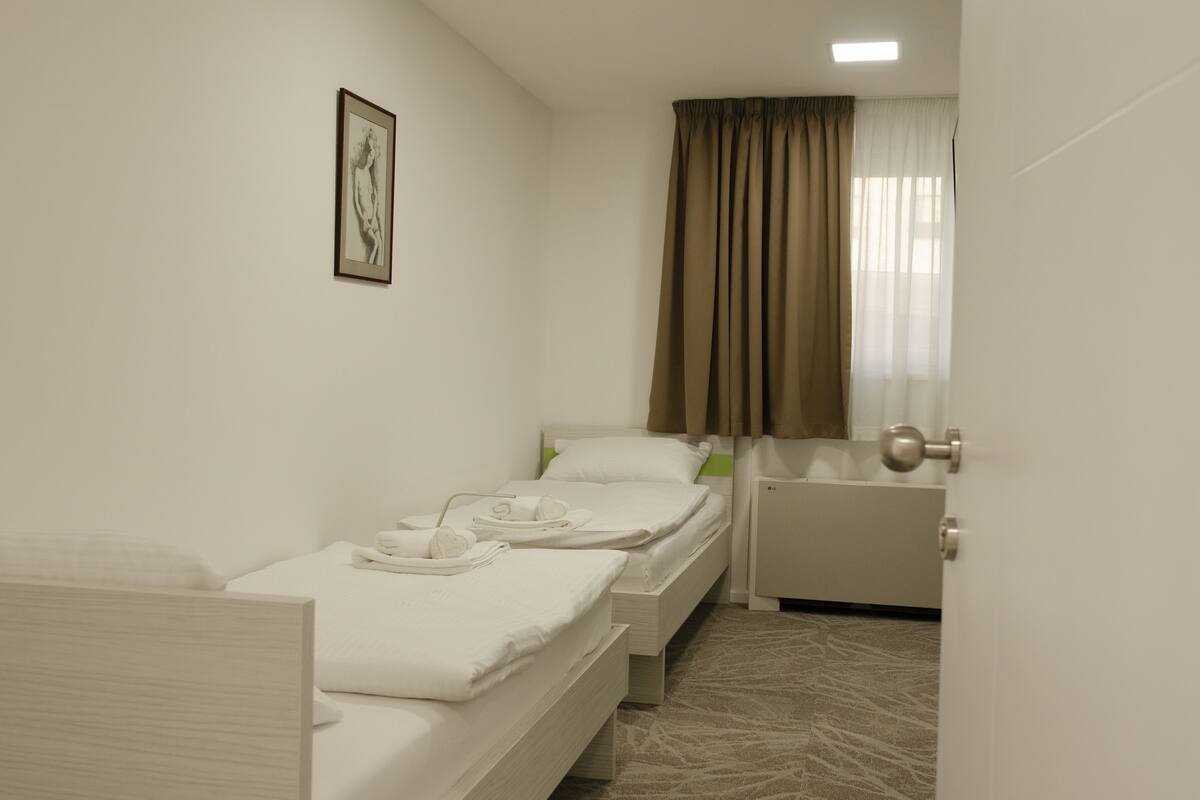 A hotel room with two single beds, a small table with a chair, a radiator, and a window with curtains.