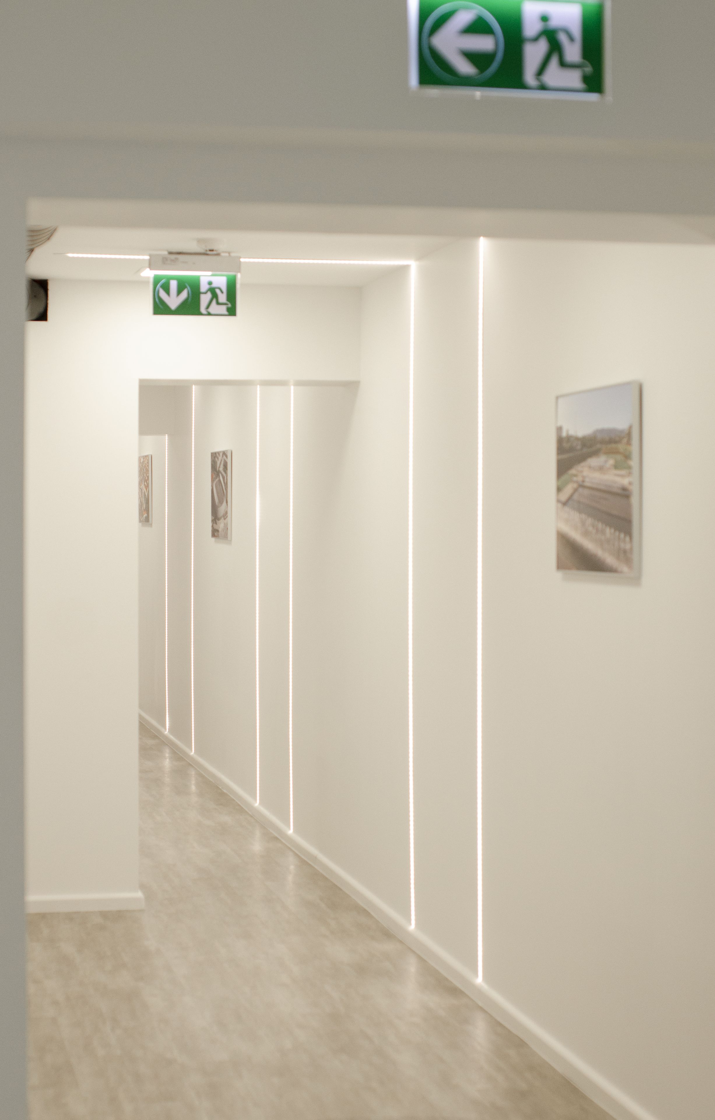 A hallway with a green emergency exit sign, a framed picture on the wall, and a light strip running along the ceiling.