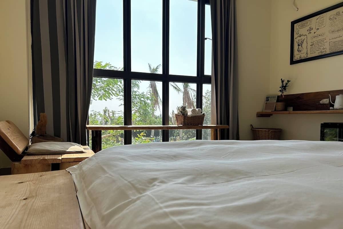 Une chambre avec un grand lit, une chaise en bois, une fenêtre donnant sur un paysage verdoyant et une étagère avec divers objets.