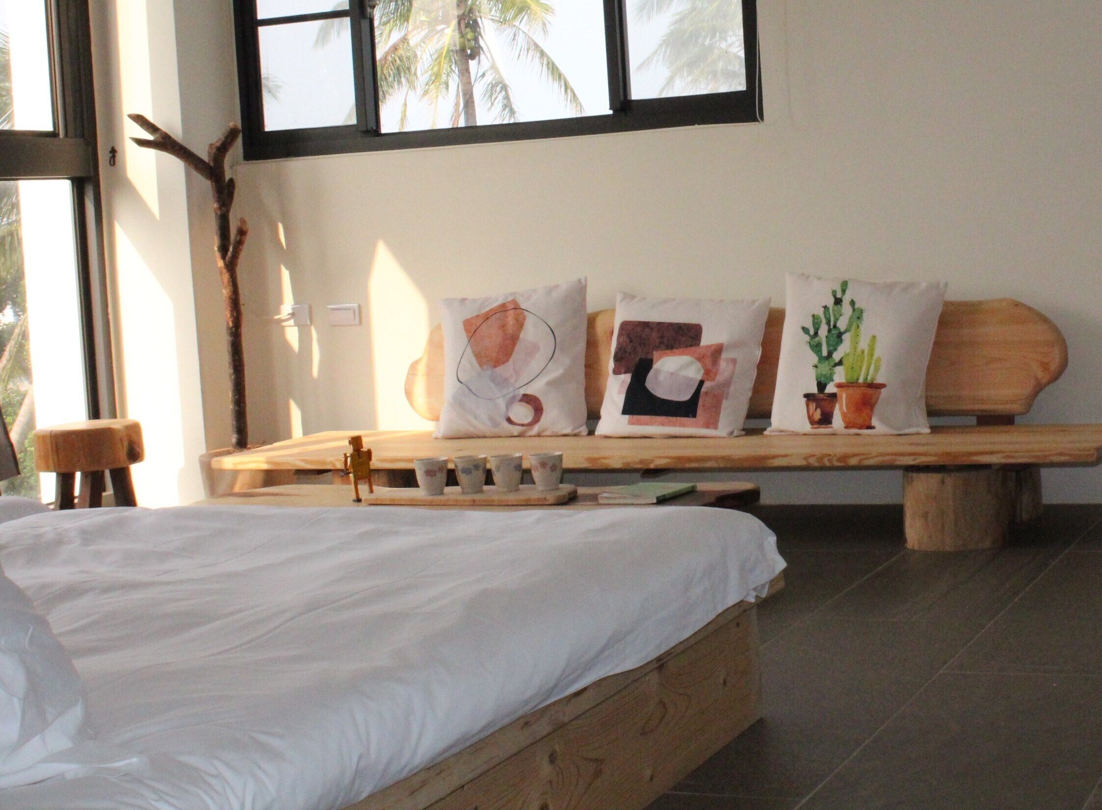 Um quarto com uma cama de madeira, um banco com almofadas, uma mesinha com xícaras e uma janela com vista para palmeiras.