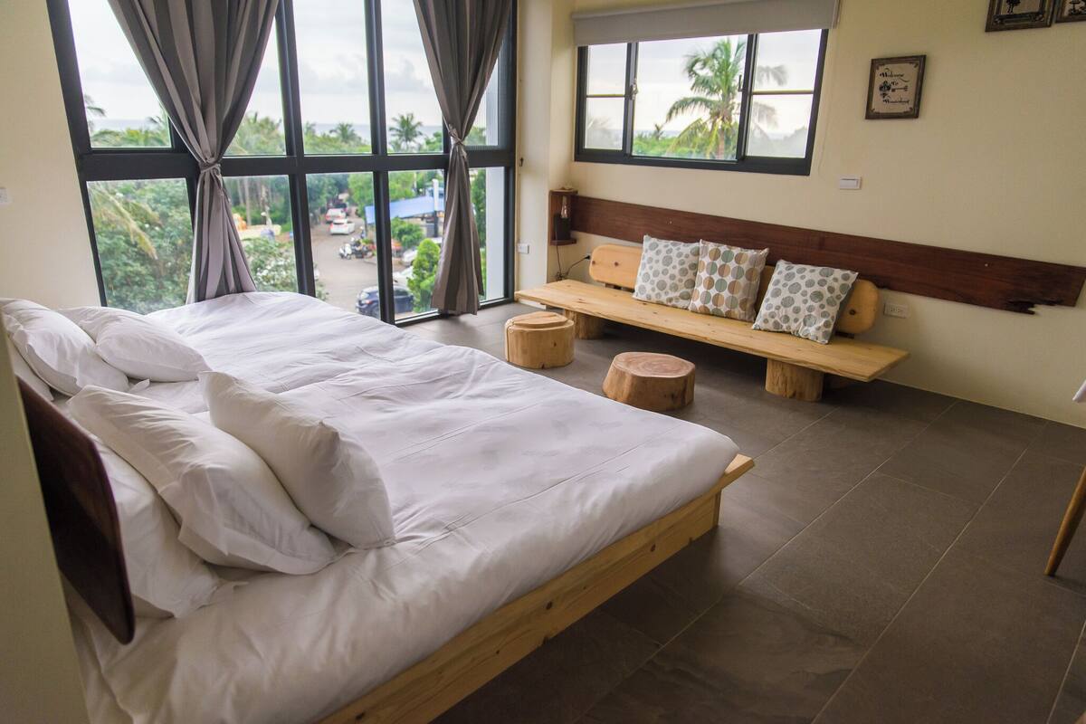 Une chambre à coucher avec un grand lit, une tête de lit en bois, un banc avec des coussins et une vue sur l’extérieur.