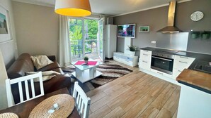 En moderne stue med sofa, sofabord og tv. Der er et stort vindue, et køkken med komfur samt et spiseområde med stole.
