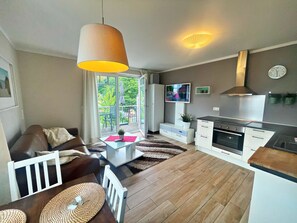 Ein modernes Wohnzimmer mit einer Couch, einem Couchtisch und einem Fernseher. Es hat ein großes Fenster, eine Küche mit Herd und einen Essbereich mit Stühlen.