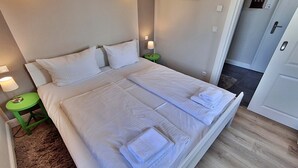 En pænt redt dobbeltseng med hvide sengetøj og håndklæder, et grønt natbord med lampe, og et billede i ramme på væggen.