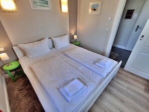 Ein ordentlich bezogenes Doppelbett mit weißen Leinen und Handtüchern, ein Nachttisch in Grün mit einer Lampe und ein gerahmtes Bild an der Wand.