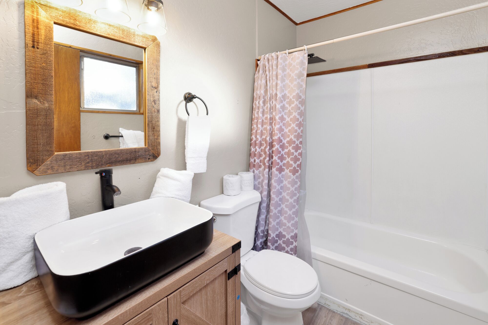 A bathroom with a wooden vanity, a black sink, a white toilet, a shower with a patterned curtain, and a mirror with a wooden frame.