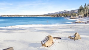 A snowy beach with clear blue water, a few rocks, and a forested mountain in the background. - Shore Acres Lodge #452 (Big Bear Lake)