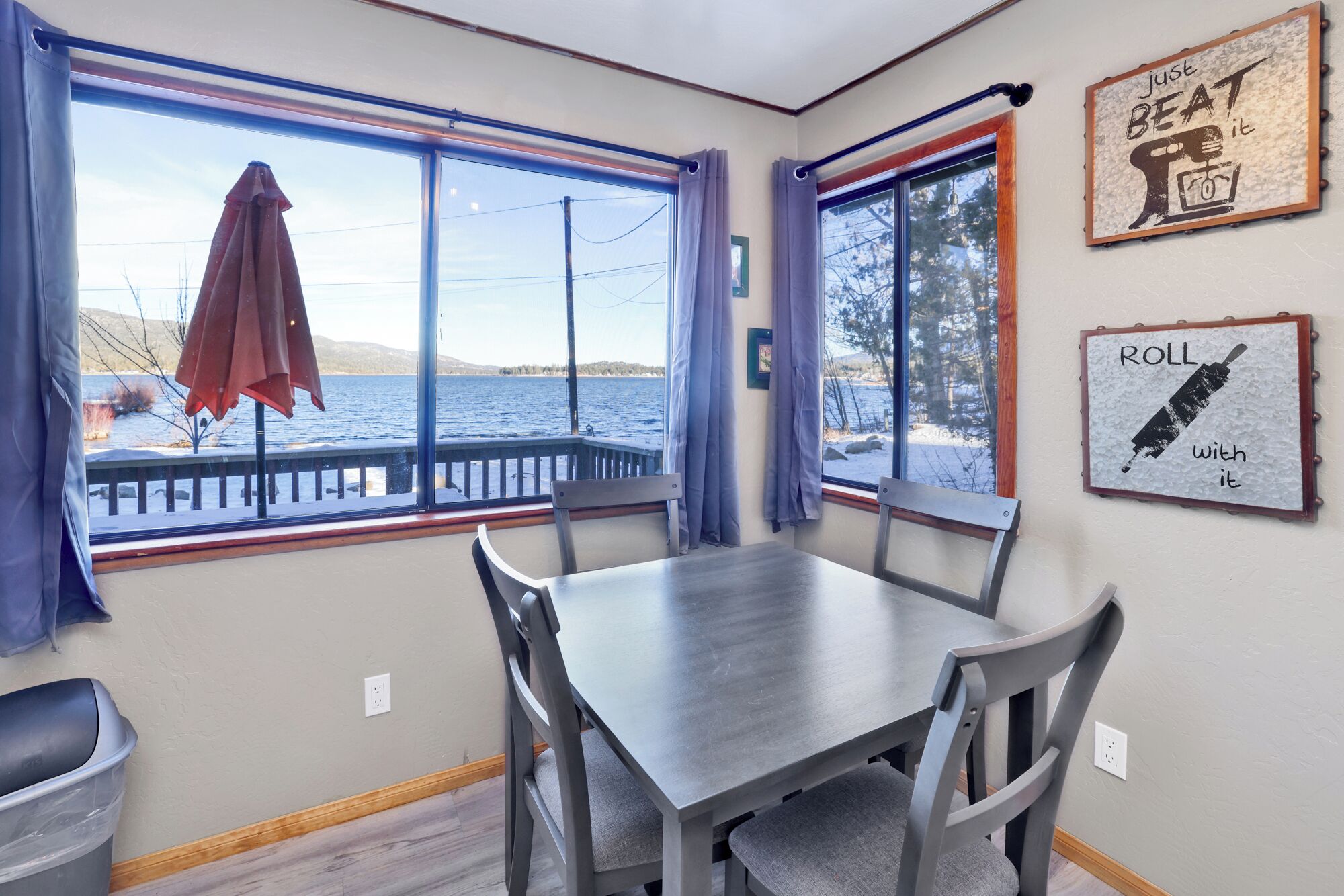 A dining area with a table and chairs, large windows with curtains, and two wall decorations.