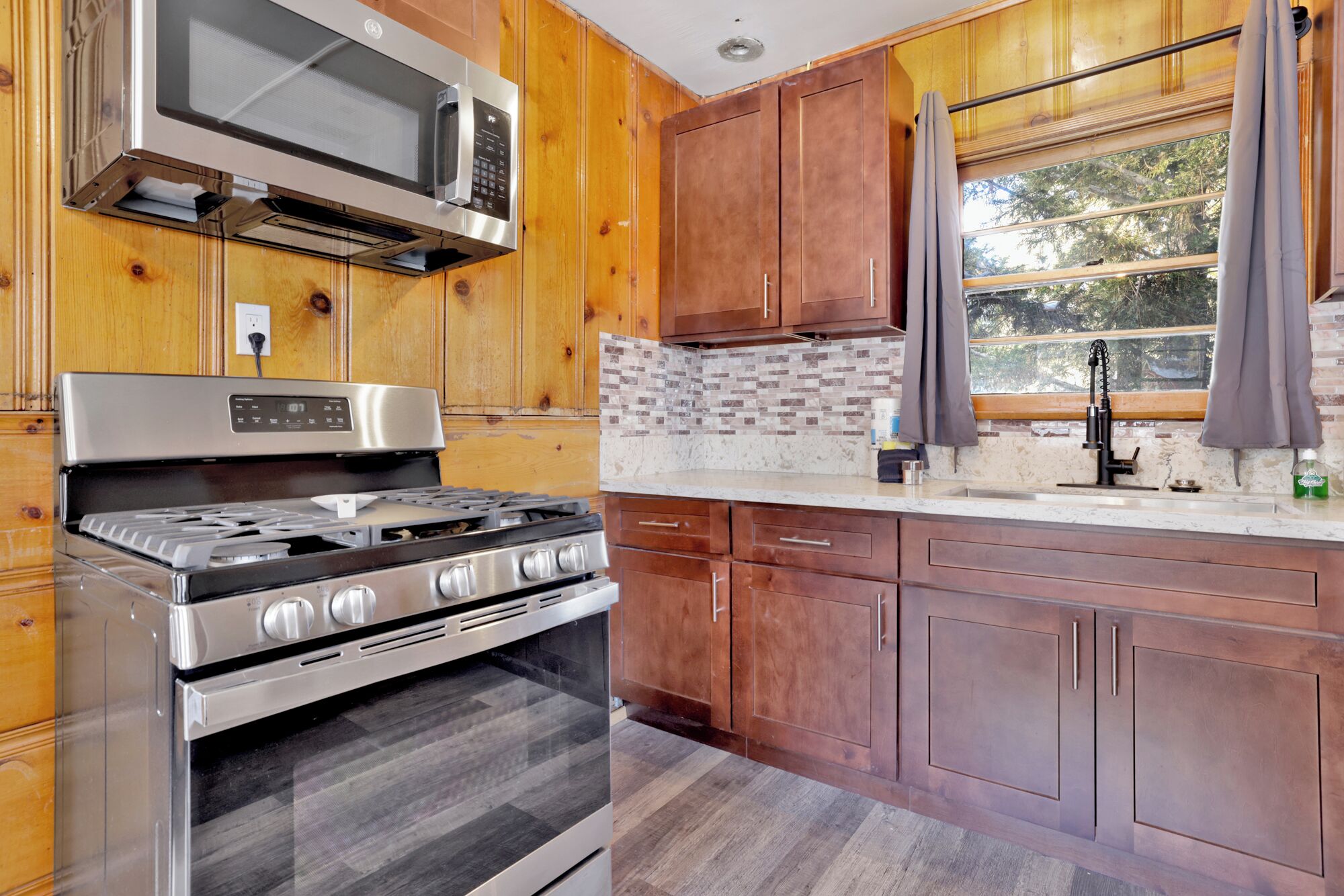 A kitchen with wooden cabinets, a stainless steel stove, a microwave, and a window with curtains.