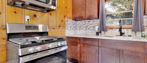 A kitchen with wooden cabinets, a stainless steel stove, a microwave, and a window with curtains.