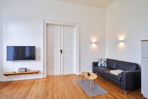 A modern living room with a flat-screen TV, a wooden shelf, a black leather sofa, and a small wooden table.