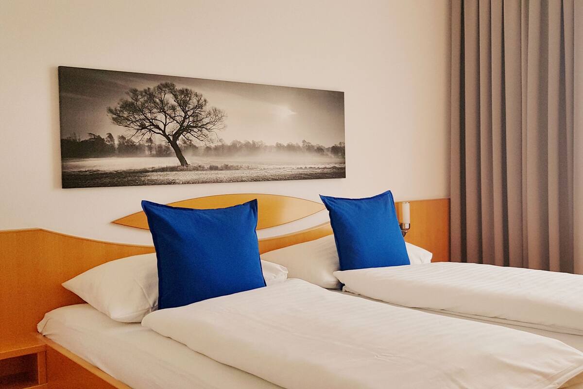 A hotel room with a bed, two pillows, a nightstand, a lamp, a window with curtains, and a painting on the wall.