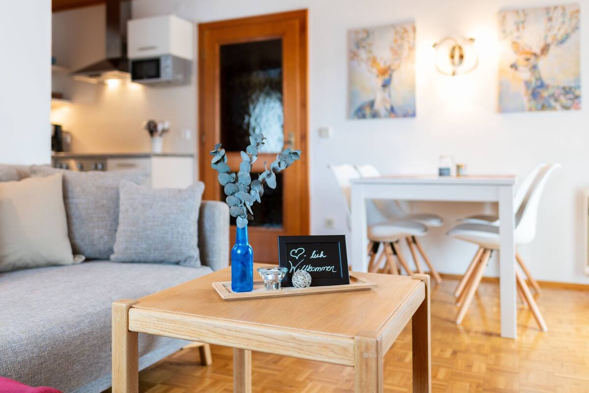 A living room with a sofa, a small wooden table with a vase and a welcome sign, a dining area with a white table and chairs, and a kitchen in the background.