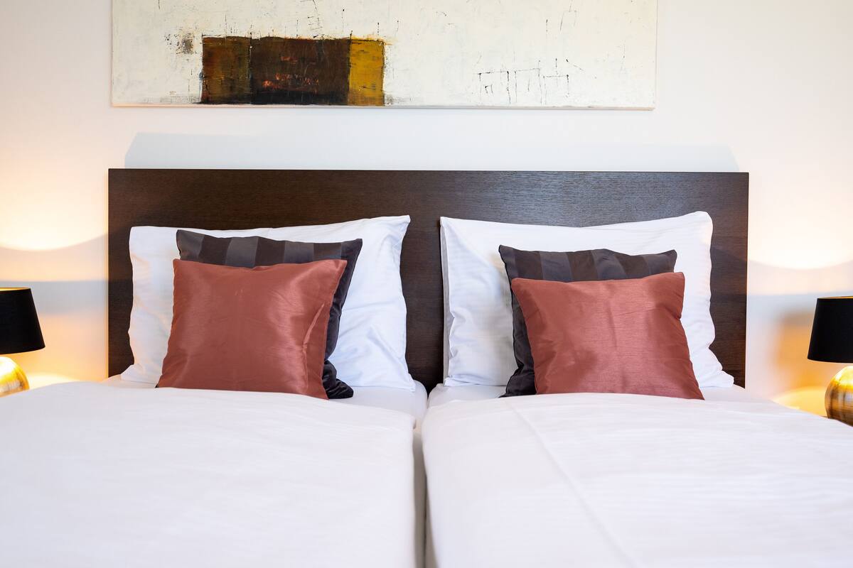 A double bed with white bedding and two brown pillows, flanked by two bedside lamps.