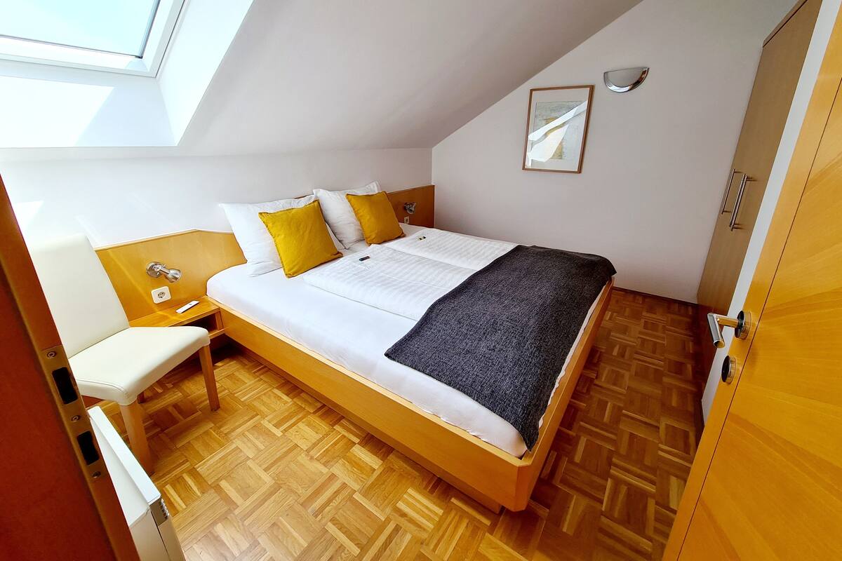 A bedroom with a bed, a bedside table, a chair, a window, and a wooden floor.