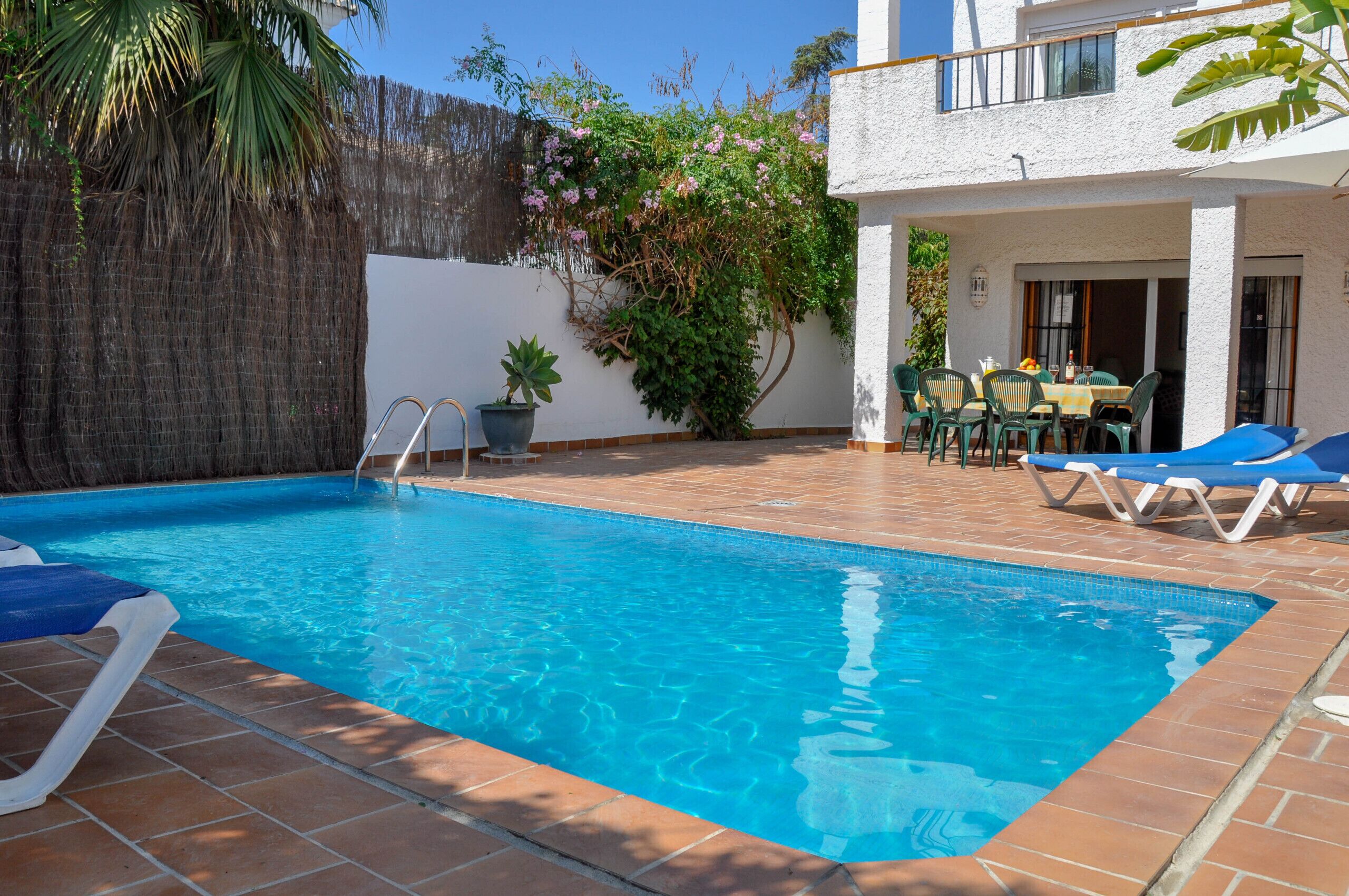 A rectangular swimming pool with clear blue water, surrounded by a tiled patio. There are lounge chairs and a table with chairs outside the pool area.