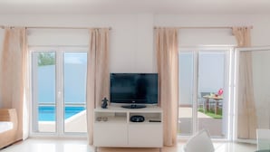 Una sala moderna con un televisor de pantalla plana sobre un mueble blanco, una mesa de vidrio y vistas a una piscina y un área de estar al aire libre.
