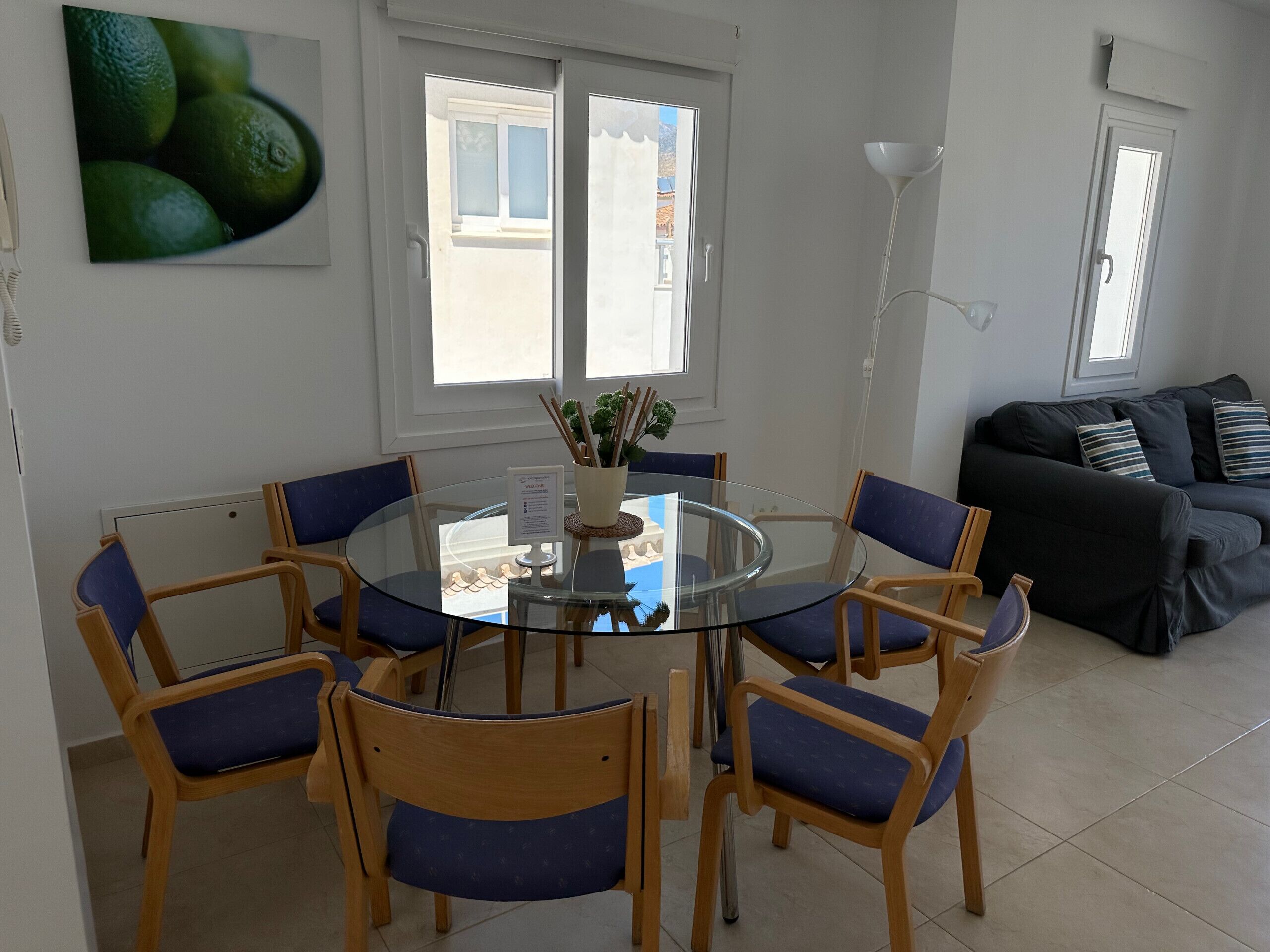 A dining area with a round glass table surrounded by wooden chairs with blue cushions, a black sofa with striped cushions, and a window with a view of buildings.