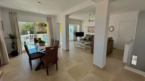 Un'ampia zona giorno con tavolo da pranzo, sedie, divano e televisore. Include un balcone con vista sul verde.