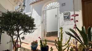 Un edificio bianco con tetto in tegole, una targa con il numero 17 e una porta con finestra. In primo piano, piante in vaso e un cactus.