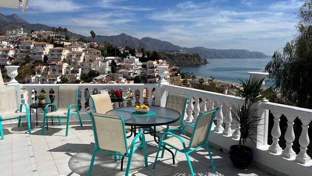 Una terrazza con sedie color turchese e un tavolo, affacciata su una cittadina costiera con edifici bianchi e vista sul mare.