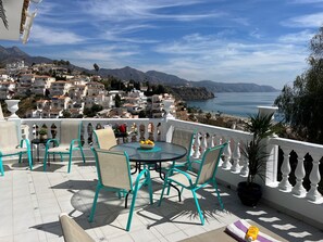 En terrasse med turkise stole og et bord, med udsigt over en kystby med hvide bygninger og havudsigt.