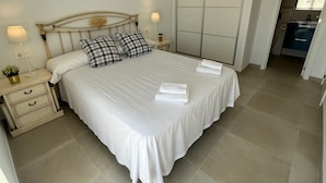 A neatly made bed with white linens and checkered pillows, a bedside table with a lamp, and a view into a bathroom. - Nerja Paradise Rentals - Villa Las Palmeras (Nerja)