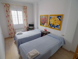 Two single beds with neatly folded towels, a radiator, and a small table with a lamp and flowers.