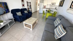 Un salon moderne avec un canapé gris, un canapé bleu, une table basse et un téléviseur. On aperçoit une cuisine au fond, avec une table à manger et des chaises.