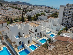 Un quartier résidentiel avec des bâtiments blancs, une piscine sur le toit avec de l’eau bleue, et une vue sur la mer et les montagnes en arrière-plan.