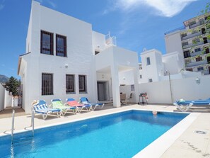 Un bâtiment moderne de couleur blanche, doté d’une piscine, de chaises longues et surplombant un ciel d’un bleu limpide.