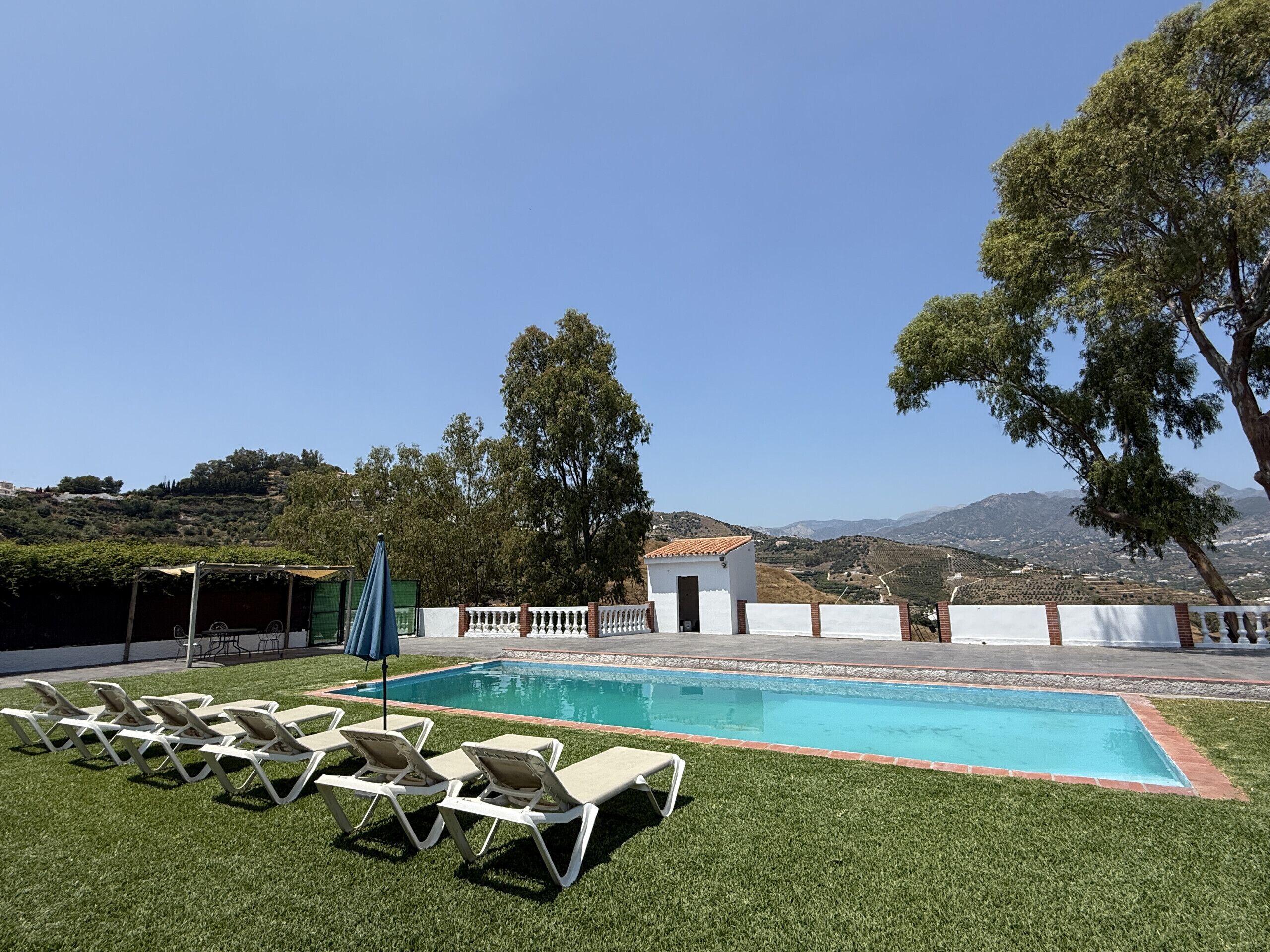 A swimming pool with lounge chairs, a small building, and a view of hills and mountains.