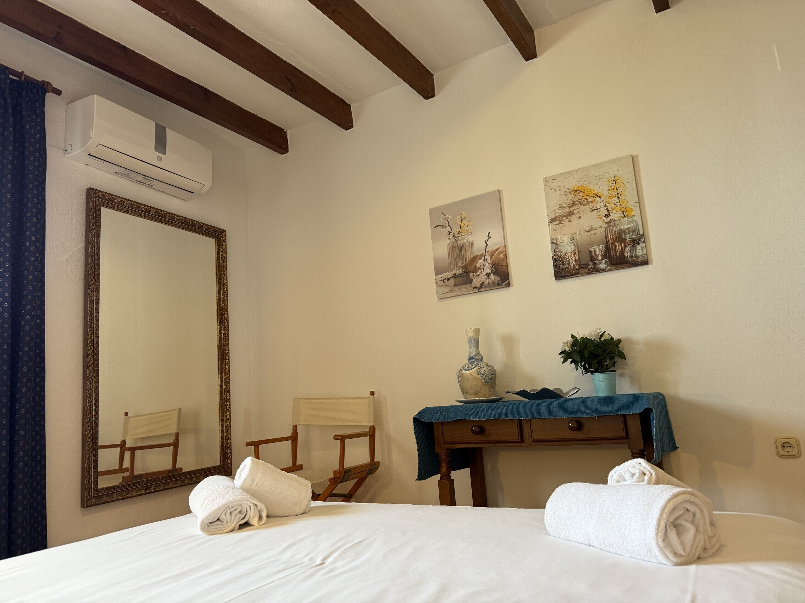 Quarto de hotel com um espelho grande, duas pinturas, uma cadeira de madeira, uma mesinha com um vaso e planta, e toalhas na cama.