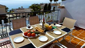 Un balcón con una mesa dispuesta para dos personas, que incluye una botella de vino, pan, tomates y una cesta de fruta.
