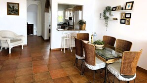 Amplia sala de estar con una mesa de comedor rodeada de sillas de mimbre, una cocina al fondo y un sofá blanco.