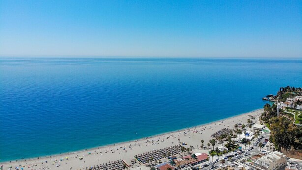 Escena costera con playa, cielo despejado, arena y un pueblo pequeño con edificios y palmeras.