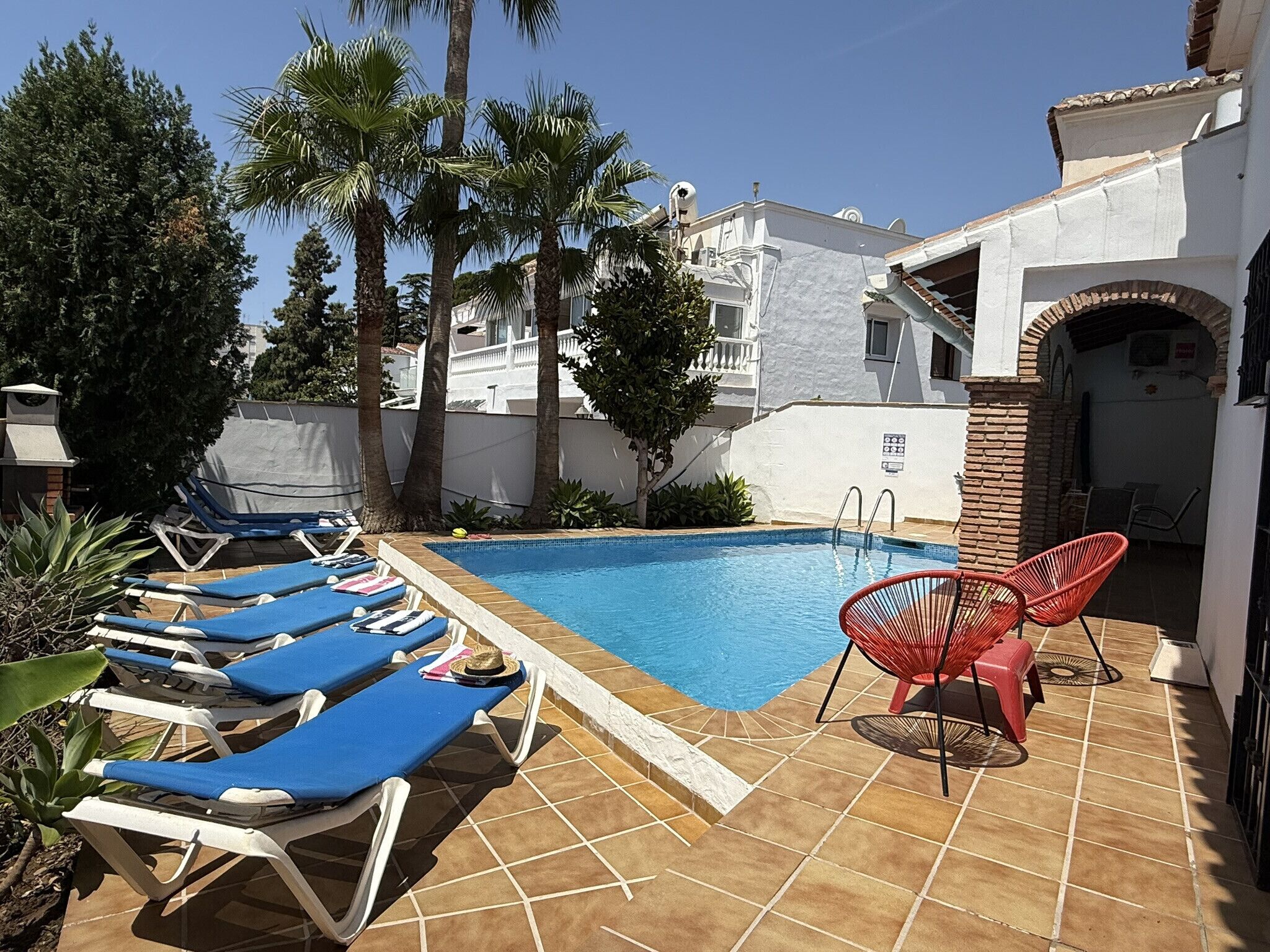 A pool area with blue lounge chairs and a red chair, surrounded by palm trees and a white building with a tiled floor.