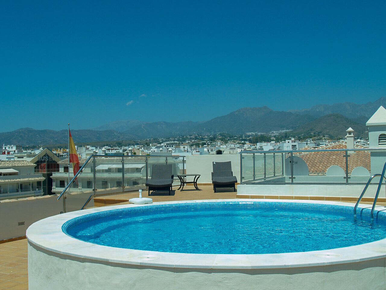A rooftop pool with two lounge chairs and a flag, surrounded by buildings and mountains in the background.