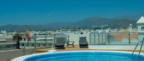 A rooftop pool with two lounge chairs and a flag, surrounded by buildings and mountains in the background.