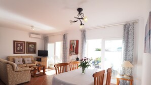 Un saló amb un sofà, televisió i taula de dinar amb caires. Hi ha una pèndol, cortines i un quadre a la paret.