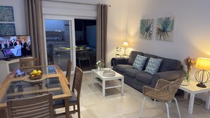 Un salon moderne avec une table à manger en verre, des chaises en bois, un canapé gris et un téléviseur diffusant une émission.