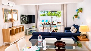 A modern living room with a glass dining table, white chairs, a blue sofa, and a TV.