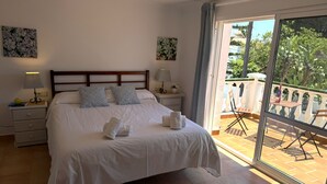 A neatly made bed with pillows and towels, a bedside table with a lamp, and a balcony with chairs and a table.